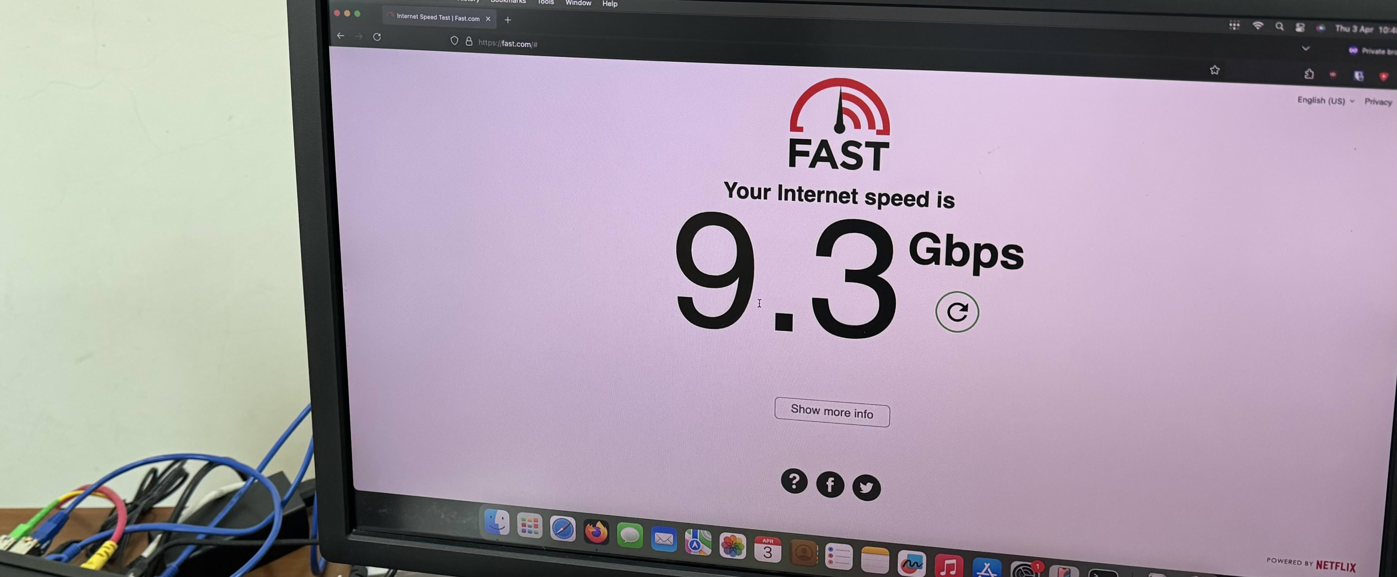 A monitor showing fast.com in Firefox with a speed test result of 9.3Gbps. On the bottom left, the router and fiber patch cord are visible